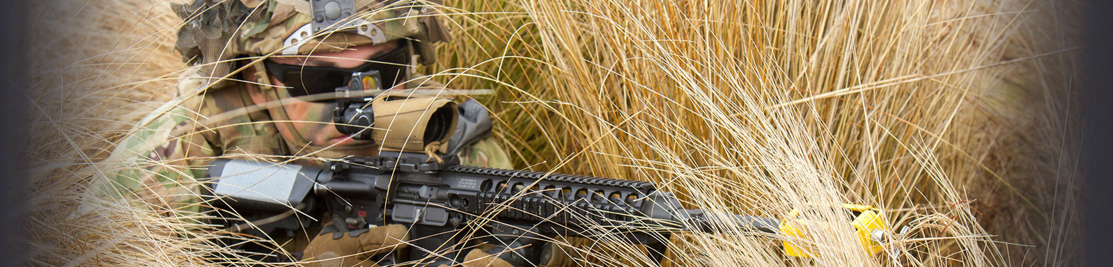 sniper laying prone in tall, dry grass.